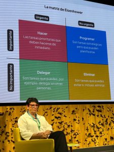 &ldquo;Diapositiva de la matriz de Eisenhower que clasifica tareas seg&uacute;n su urgencia e importancia en cuatro cuadrantes: hacer, programar, delegar y eliminar. En primer plano, una ponente sentada en una silla en el escenario durante una presentaci&oacute;n.&rdquo;